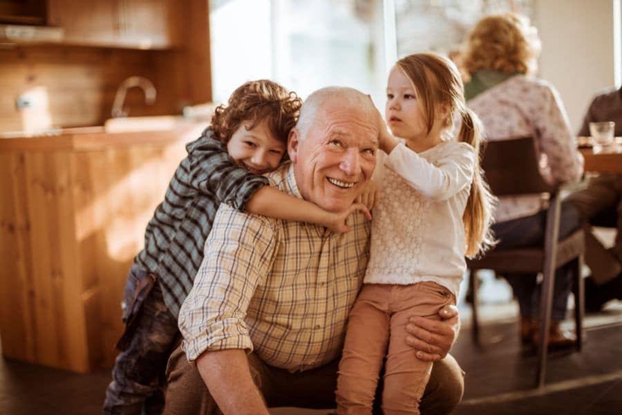 An older man playing with his grandchildren while his wife sitting in the background talking to her attorney about drafting a will.